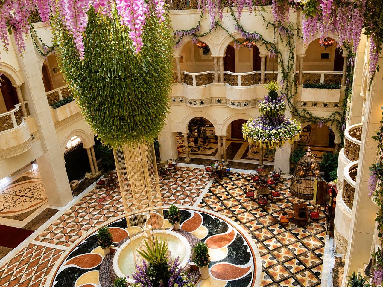 The grand multi-story atrium lobby at The Interlaken OCT Hotel Shenzhen, with carved travertine arched colonnade balconies on three floors, a geometric inlaid marble floor in red, black, gold, and cream, a central marble fountain with petal-pattern radiation, suspended cascading wisteria and floral installations, a decorative gilded bird cage, and a large hanging green topiary cone, combining Italian-Mediterranean and Khajuraho-influenced design at the OCT East resort in Yantian District